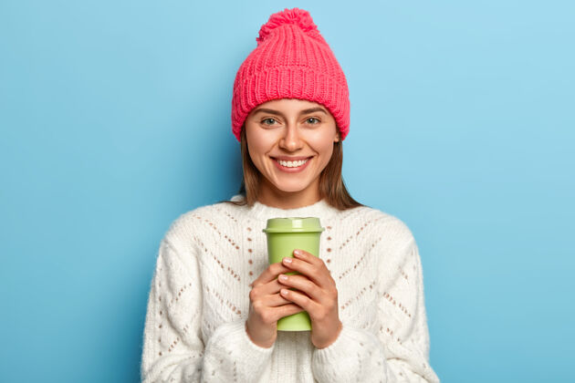 帽子快乐的女人戴着亮粉色的帽子 穿着白色暖和的毛衣 手里拿着一杯外卖咖啡 对着蓝色的墙壁摆姿势咖啡女性嘴唇