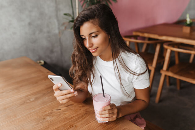 人卷曲的黑发女人穿着白色t恤在手机里写信息 坐在咖啡馆里拿着奶昔裤子奶昔晒黑