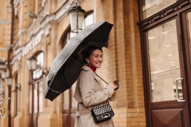 成人身着米色风衣 背着十字形包 面带微笑的美女在欧洲城市的雨伞下漫步美丽外套肖像