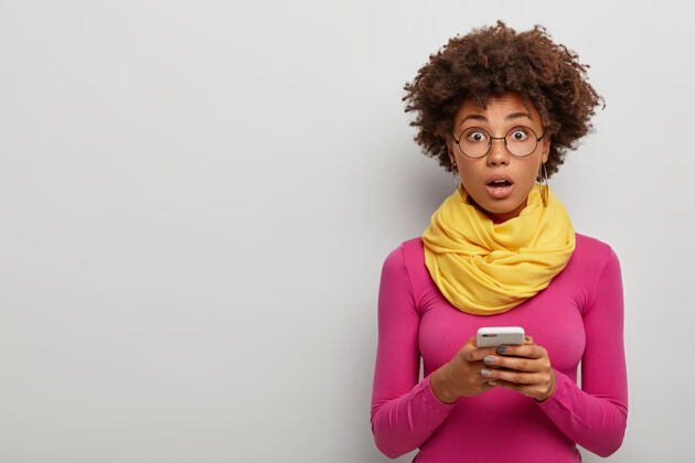 卷发震惊的非洲女人张嘴 站在室内无语 用手机在社交网络上聊天尴尬震惊随意