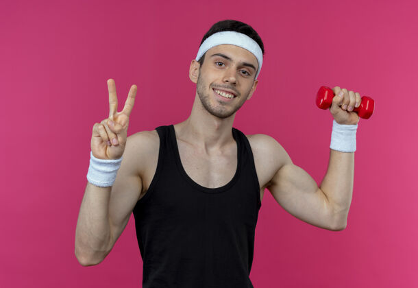 标志戴着头巾的年轻运动型男子正在练习哑铃微笑着展示战胜粉色的胜利标志粉红色胜利哑铃