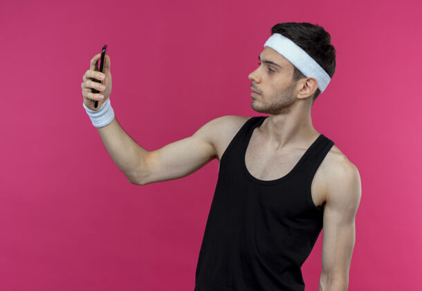 自拍戴着头巾的年轻运动型男子用智能手机在粉色外自拍年轻站立使用