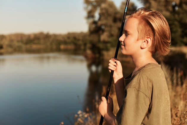 卷轴年轻的欧洲金发男孩在夏日阳光明媚的日子里钓鱼 遥望远方 享受闲暇时光 穿着绿色t恤 站在河边的水边湖泊河流运动