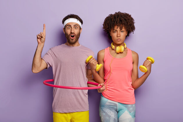 一起严肃的女人举着黄色的哑铃 穿着粉色上衣和紧身裤 惊讶的没刮胡子的男人指着上面的空白处 用呼啦圈来保持健康 孤立在紫色的墙上姿势健康锻炼