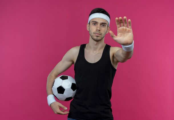 球戴着头巾 手持足球 手张开 表情严肃的年轻人站在粉红色的墙上做停车标志运动开男人