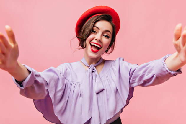 波浪发穿着红色贝雷帽和紫色上衣的时髦女人微笑着在粉色背景上自拍卷发连衣裙贝雷帽