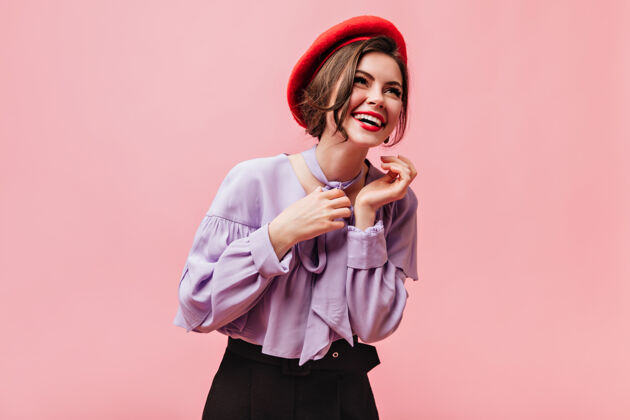 脸穿着红色贝雷帽和淡紫色上衣的积极的女人在粉色背景下大笑女性贝雷帽波浪发