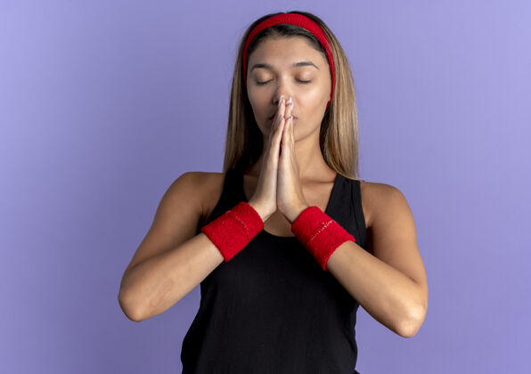 红色身穿黑色运动服 头戴红色头巾的年轻健身女孩 双手合十 带着希望的表情 闭上眼睛 祈祷着蓝色蓝色举行希望