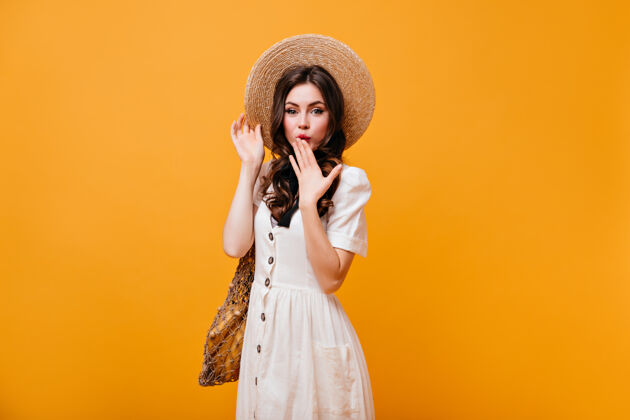 肖像绿眼睛的女士用手捂住嘴戴草帽和白色太阳裙的女士拿着橙色背景的购物袋头发脸年轻人