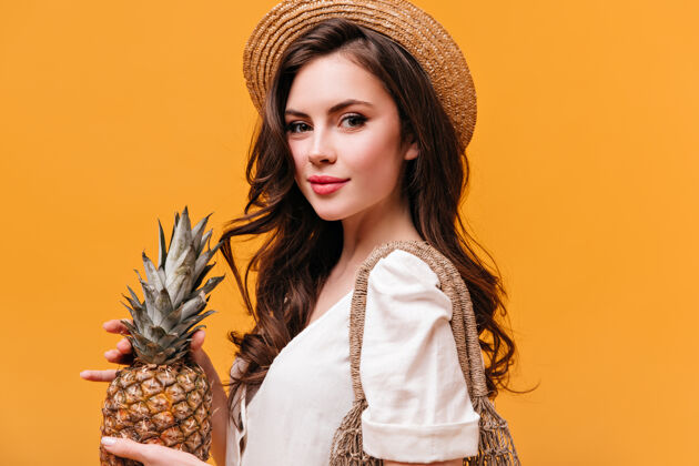 脸迷人的绿眼黑发女人 戴着草帽 手里拿着菠萝 看着橙色背景下的相机线袋肖像成人