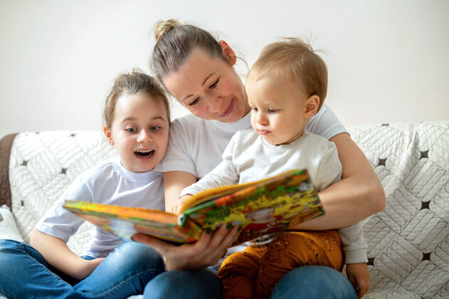 兄弟姐妹妈妈和她的两个孩子在家里的沙发上看书快乐而微笑学习年轻女人