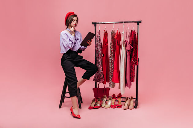 姿势穿着时髦服装和红色贝雷帽的女孩坐在椅子上 穿着闪亮的衣服和鞋子 在粉色背景上若有所思地阅读女性购物肖像