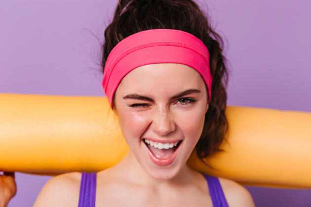 模特积极的女运动员在粉红色的头带微笑和眨眼紫色的墙壁与橙色的健身垫80年代80年代风格身材