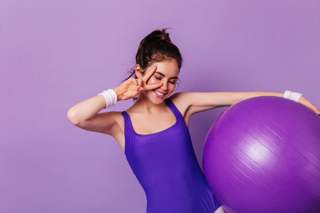 紧身裤苗条的体操女子手持健身球 在紫色的墙上显示和平标志健康健美操时尚