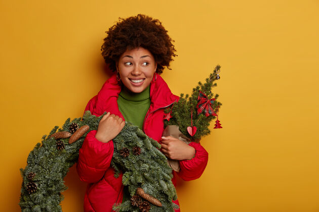 枞树迷人的卷发女人咧嘴笑着 看向一边 穿着外套 手里拿着装饰过的小枞树和云杉花环圣诞节云杉室内