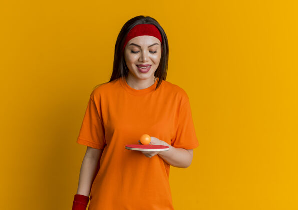 运动带着头带和腕带微笑的年轻运动女性手拿乒乓球拍看着上面有乒乓球橙色乒乓球腕带