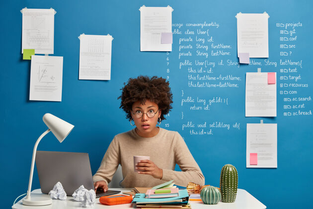 分析迷惑不解惊讶的电脑分析师喝咖啡休息 阅读网页上发表的文章 戴着圆眼镜 对着蓝色的墙壁在桌面上摆姿势 上面写着信息桌子办公桌纸张