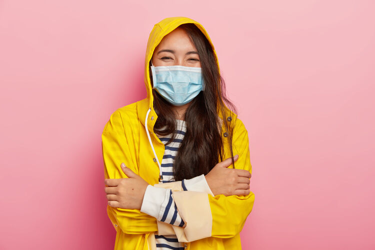 疾病快乐美丽的黑发女子 双臂合十 戴着医用口罩 穿着防水雨衣 抵御季节性疾病天气雨衣面具