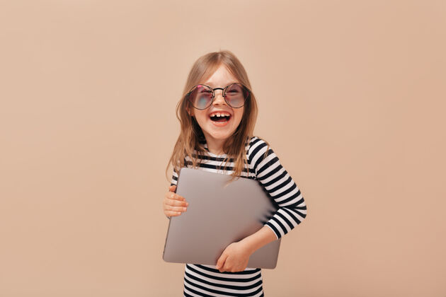 年轻摄影棚拍摄的微笑快乐可爱的女孩戴着时髦的眼镜 拿着笔记本电脑在米色背景下太阳镜孩子享受