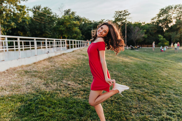 草幸福的红衣女孩在草地上滑稽地跳舞在温暖的日子里 优雅的黑发女人在公园里拍照时大笑年轻休闲放松