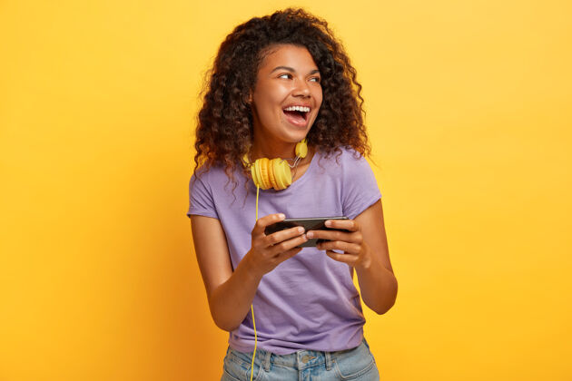 请高兴的卷发女人与非洲发型 手持智能手机水平 玩游戏在线无线年轻欣喜若狂