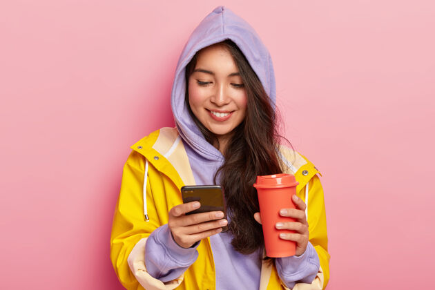 信息快乐的黑发女孩戴着兜帽和雨衣 拿着手机 在社交网络上滚动 喝咖啡或茶快乐电话女性