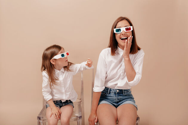 黑发母亲和女儿面带微笑地看着彼此 戴着3d眼镜在米色背景的沙发上对着摄像机微笑快乐女性