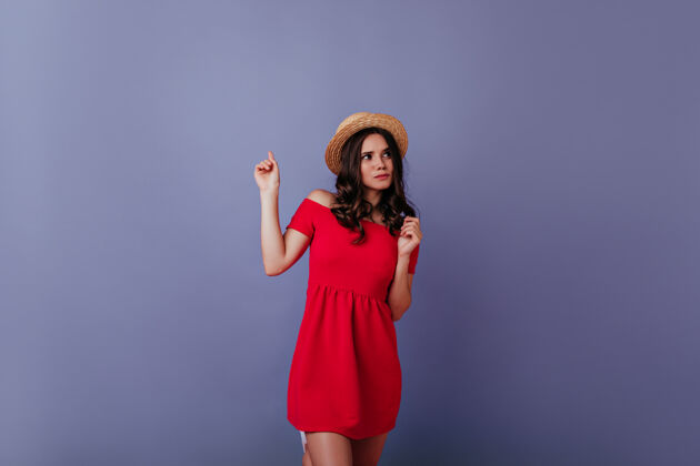 优雅一个穿着考究的女孩在紫色的墙上摆着姿势一个漂亮的黑发女人穿着红色的裙子 戴着草帽 享受着拍照的乐趣享受成人积极