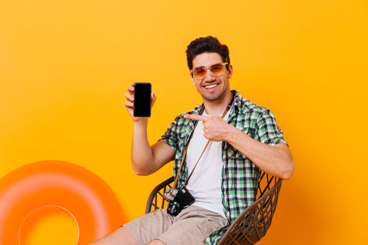 休闲夏日行头的迷人男人指着黑色智能手机欢快的家伙拿着复古相机坐在椅子上 橙色的空间里放着充气圈冒险啤酒男孩