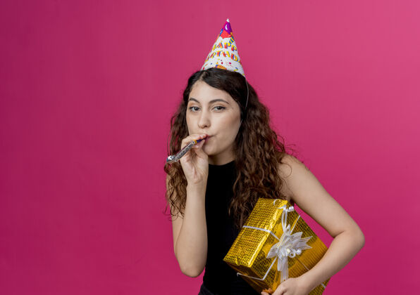 卷发年轻漂亮的女人 卷发 戴着节日帽吹着口哨 手里拿着礼盒 站在粉色的墙上 生日派对的概念吹口哨年轻吹