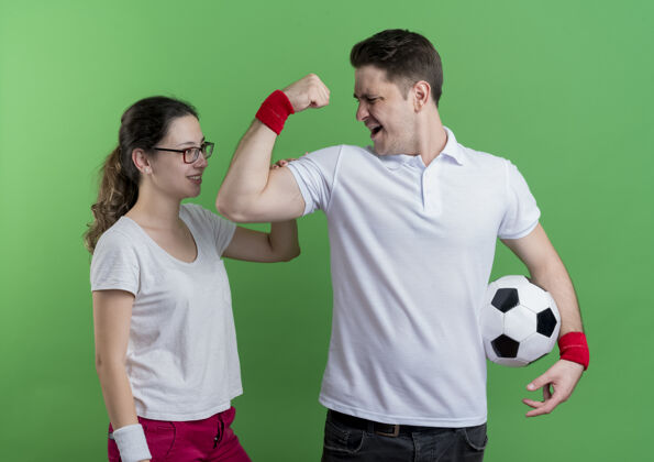 二头肌一对年轻的运动型情侣站在他微笑着的女友旁边 手拿足球 二头肌在绿色的墙上足球微笑球