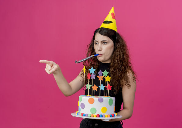 帽子一个年轻漂亮的女人 卷发 戴着节日帽 手里拿着生日蛋糕吹着口哨 手指指向一边 站在粉色的墙上 看起来很不高兴的生日派对概念侧身年轻看