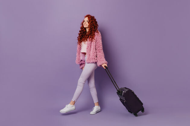 酷穿着粉色外套和白色牛仔裤的红发旅行者女孩在紫色空间带着手提箱手提箱优雅裤子