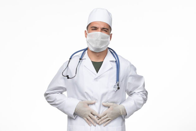 正面正面图男医生穿着医疗服 戴着口罩 在白墙上抵御胃痛冠状病毒-冠状病毒病隔离大流行观点医生面具