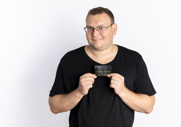 穿戴眼镜的超重男子身穿黑色t恤展示信用卡站在白墙上看着摄像机 脸上露出幸福的表情男人超重立场