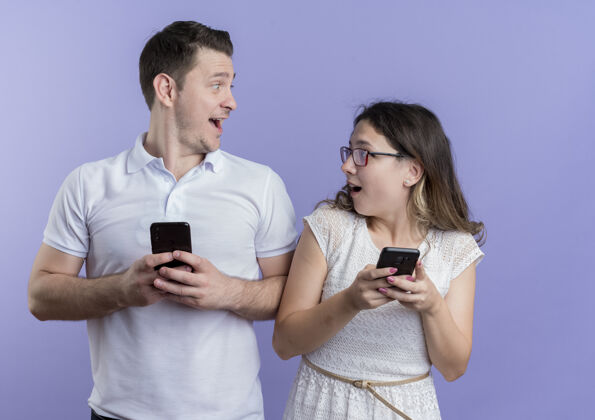 年轻一对年轻夫妇手持智能手机 站在蓝色的墙上 惊讶而快乐地看着对方情侣移动男人