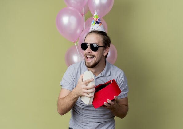 庆祝戴着节日帽的年轻人举着礼盒庆祝生日快乐而兴奋地微笑着 手里拿着气球站在轻墙上举行欢呼光