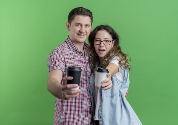 情侣一对穿着休闲服的年轻夫妇站在绿色的墙上 展示着咖啡杯快乐而积极的微笑年轻积极衣服