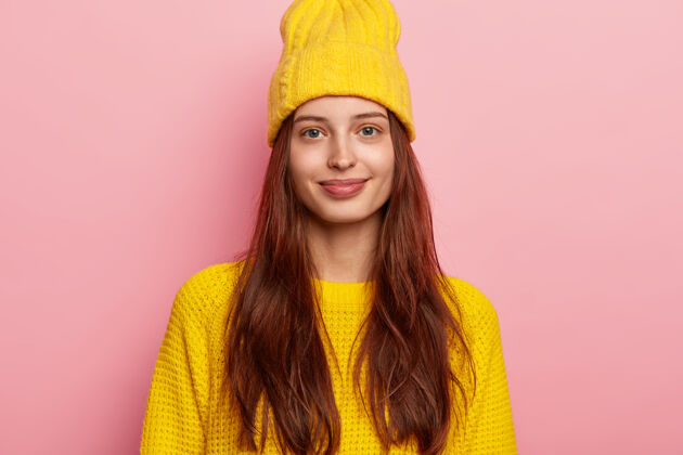 毛衣一张漂亮的女模特的照片 留着长长的黑发 端庄地看着镜头 戴着鲜艳的黄色帽子和针织毛衣 心情很好 隔离在粉色的背景下编织一个享受