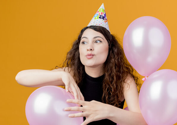 帽子一位年轻漂亮的女士 卷发 戴着节日帽 手持气球 站在橙色的墙上 快乐而兴奋地庆祝生日卷发女人站着