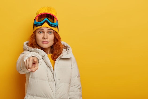 红发尴尬的姜女直指镜头 有着惊讶的表情 穿着暖和的衣服 在滑雪场 有冬天的旅游 孤立的黄色背景 有自由的空间印象外套奇迹