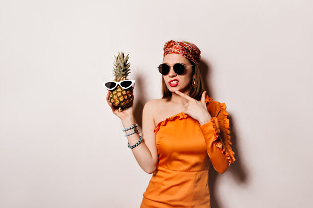 酷一位身着橘色长裙 戴着墨镜的女士若有所思地摆着姿势 手里拿着菠萝站在与世隔绝的空间里头发女士心