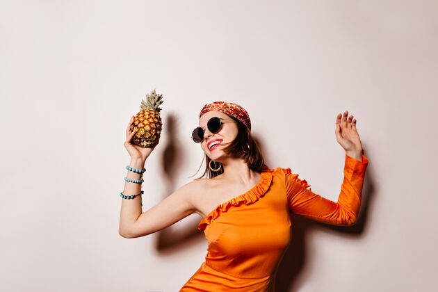 明亮身着橙色服装的女士笑着 跳舞 在白色空间里拿着菠萝太阳镜华丽女士