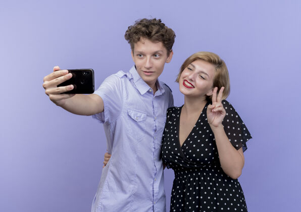 采取年轻漂亮的夫妇站在一起用手机自拍微笑着在蓝色的墙上展示v标志在一起手机手机