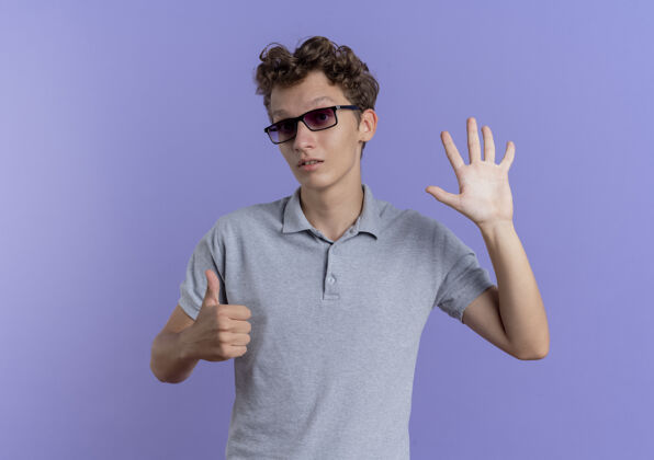站戴着黑眼镜 穿着灰色马球衫的年轻人站在蓝色的墙上 用手指指着上面男人手指六