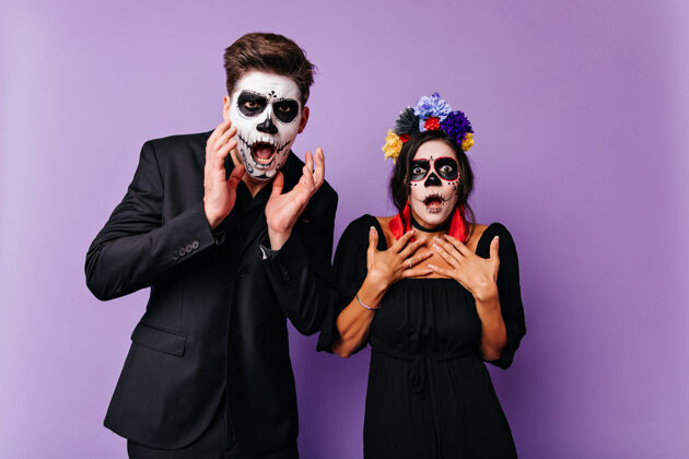 墨西哥震惊的黑发白人妇女穿着万圣节服装摆姿势惊讶的僵尸男人和女友站在紫色的墙上传统年轻花