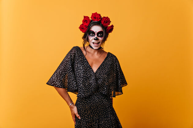 传统带着墨西哥骷髅面具的微笑女孩头发上插着玫瑰的模特在橙色的墙上笑着可怕万圣节新娘