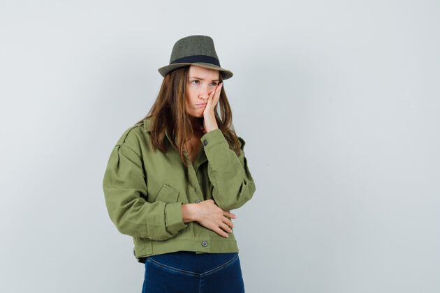 成人年轻女性穿着夹克 裤子 帽子 手放在脸颊上 神情沮丧 前视帽子黑发时尚