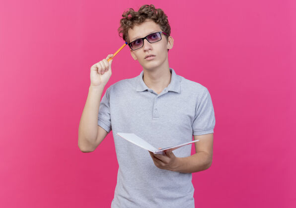 马球戴着黑眼镜的年轻人 穿着灰色马球衫 手里拿着笔记本 手里拿着笔 神情沉思 站在粉色的墙上思考拿着男人眼镜