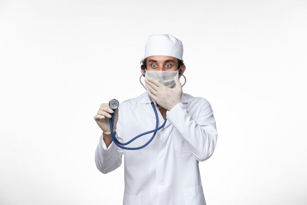 套装正面图：男医生穿着医疗服 戴着口罩以防病毒感染-使用听诊器对白色桌面病毒疾病进行病毒感染-大流行防护冠状病毒预防外套